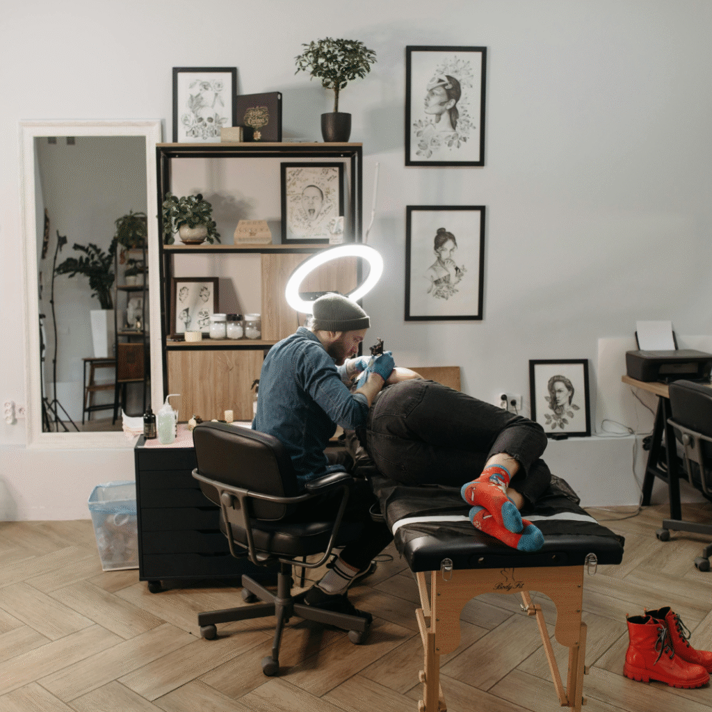 Tattoo artist working on a client’s arm in a modern studio with framed artwork on the wall and a ring light overhead.