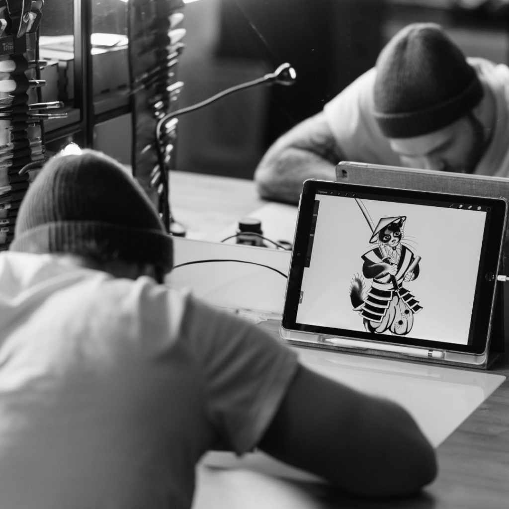 Tattoo artist sketching at a desk with a tablet displaying a samurai cat tattoo design.