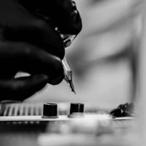 Close-up black-and-white shot of a gloved tattoo artist holding a needle above two ink caps.