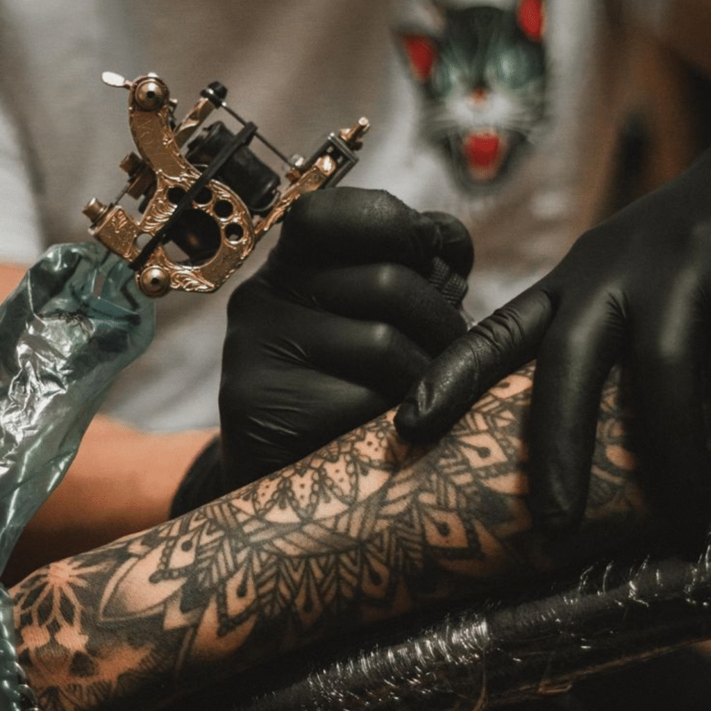 Close-up of a tattoo artist in black gloves using a brass tattoo machine to shade a mandala design on a client’s forearm.