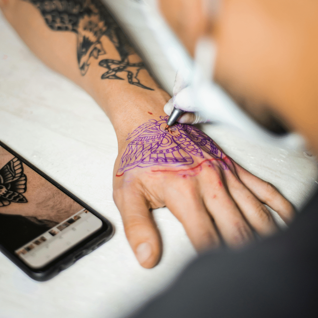 Tattoo artist outlining a hand tattoo using a purple stencil while referencing a design on a smartphone.