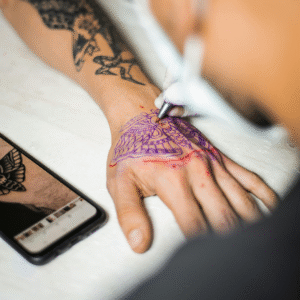 Tattoo artist outlining a hand tattoo using a purple stencil while referencing a design on a smartphone.