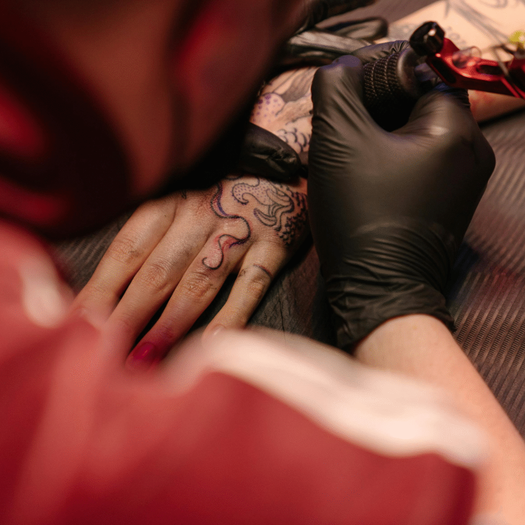 Tattoo artist in black gloves working on a detailed hand tattoo with a red rotary machine.