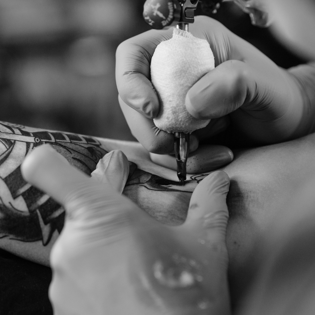 Black-and-white close-up of a gloved tattoo artist shading an arm piece with a rotary machine.