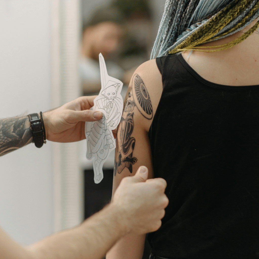 Tattoo artist holding up a samurai cat sketch next to a client’s existing shoulder tattoo to compare placement and style.
