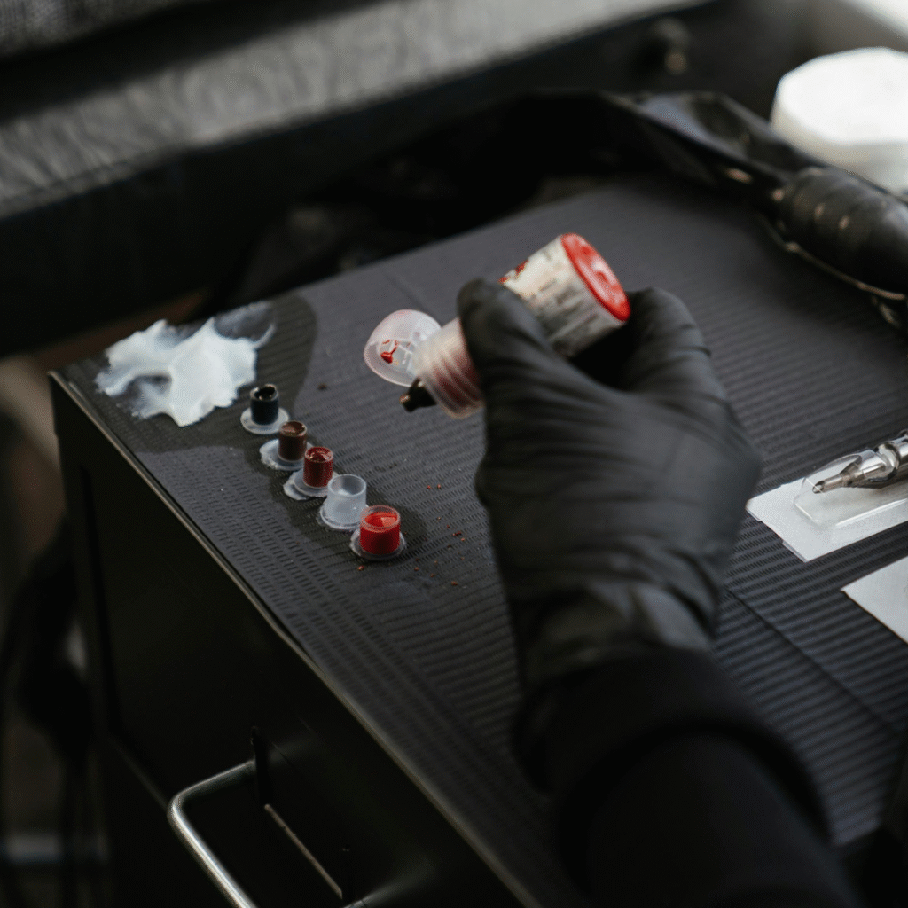 Close-up of a tattoo artist’s black-gloved hand filling small ink caps with red tattoo ink on a black work tray.