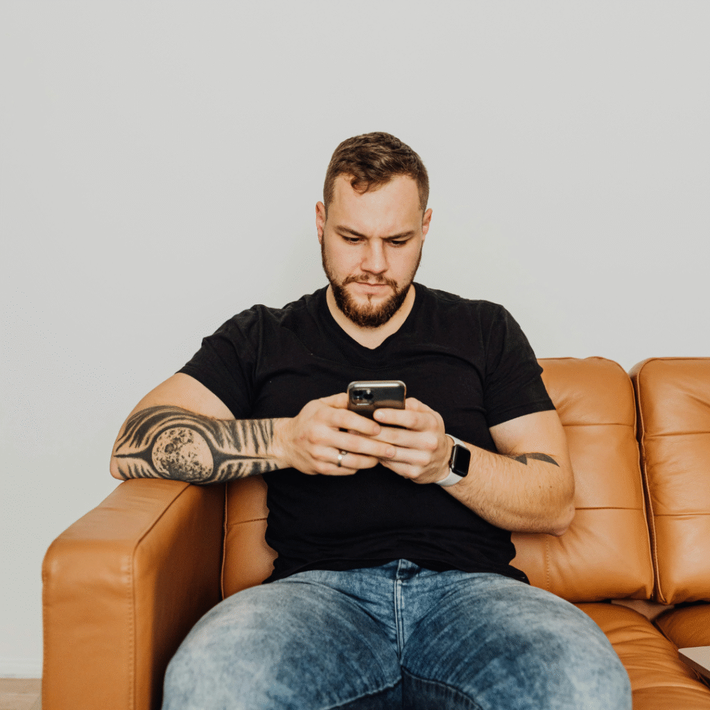 Tattooed client sitting on a couch, hesitating while looking at their phone—illustrating common tattoo booking hesitation behavior.