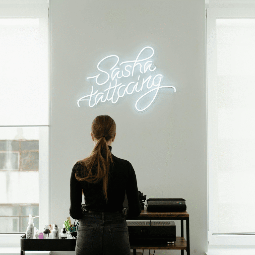 Tattoo artist standing in a bright studio beneath a neon “Sasha Tattooing” sign, representing a streamlined tattoo artist scheduling routine.