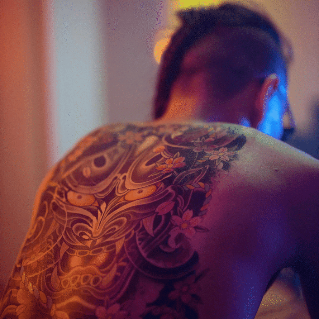 Artist’s client leaning forward, showcasing a full-back demon-mask and floral tattoo under vibrant, neon-hued studio lighting.