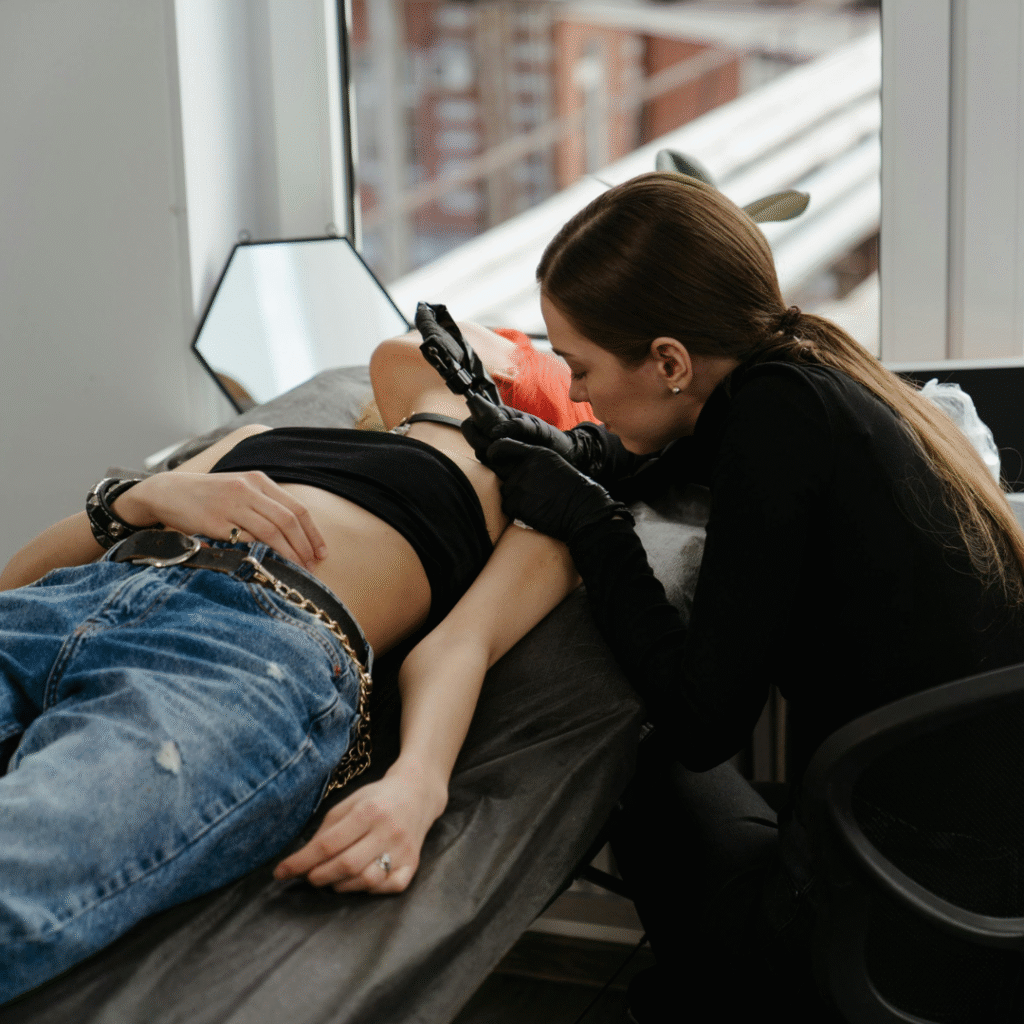 Female tattoo artist in a bright studio, wearing black gloves and carefully inking a client’s rib area.