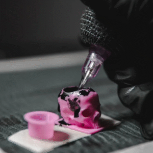Tattoo machine needle dipping into pink skull-shaped ink cap with black ink