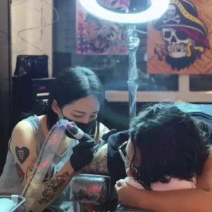 Tattoo artist focused on leg tattoo design under bright ring light reflection