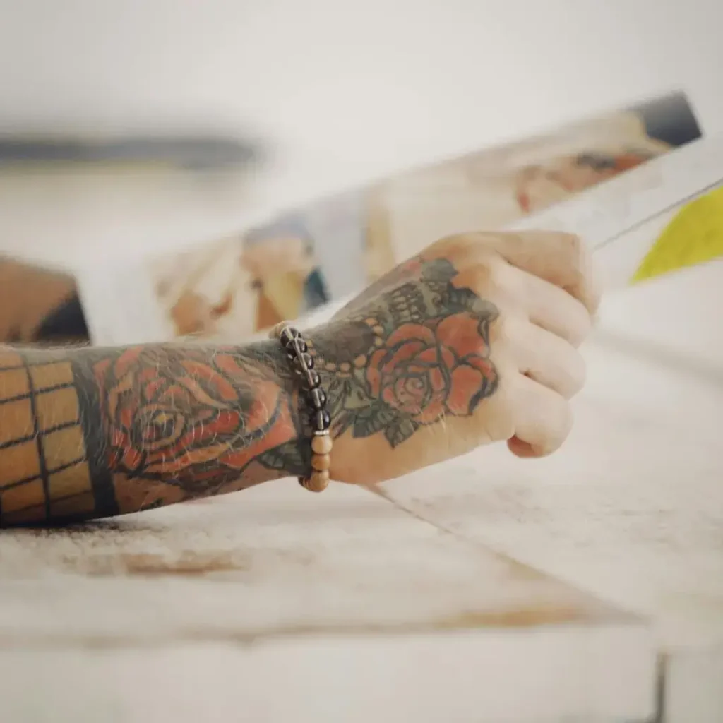 Tattooed hand with rose designs holding a magazine, representing a calm moment for artists during the holiday tattoo season.